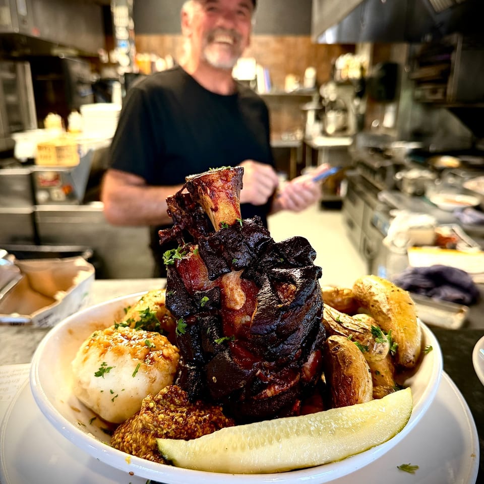 Hearty restaurant platter piled with roasted meats, potatoes, pickles, and bread, with a smiling chef working in the kitchen behind it.
